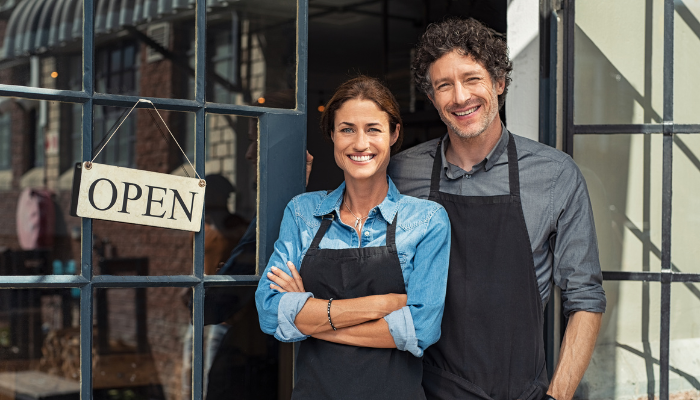 Two small business owners standing in the doorway of their shop with an open sign, smiling confidently.
