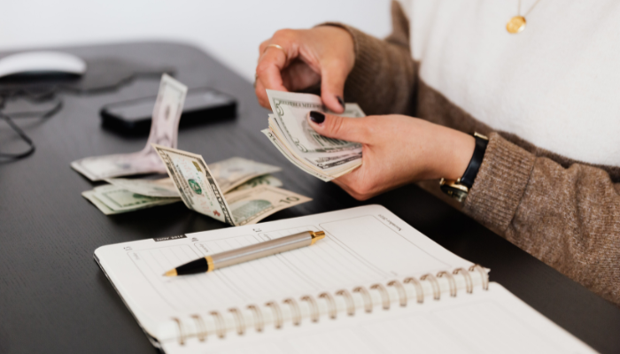 Individual counting U.S. dollar bills at a desk with a notepad and pen, managing business cash flow.