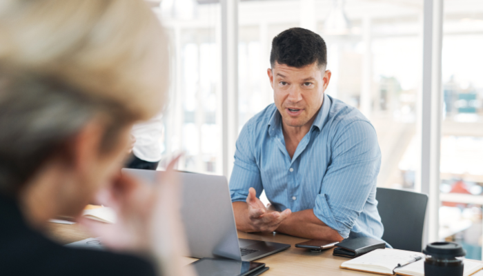 Business professional speaking to a colleague during a meeting, seated at a conference table with a laptop.