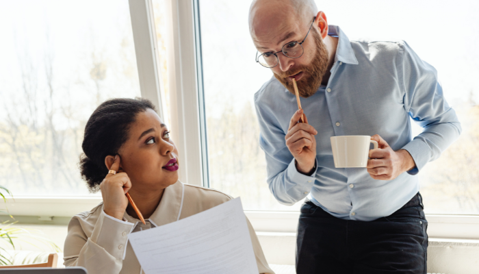 Two colleagues discussing a printed document, one holding a pencil and a coffee cup while leaning over the other’s desk.