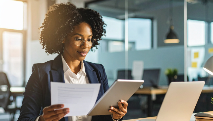 Businesswoman reviewing documents at her desk while working on a tablet and laptop in a modern office.