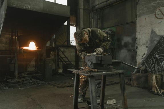 Metalworker grinding a steel component in an industrial workshop with sparks flying and a glowing furnace in the background.