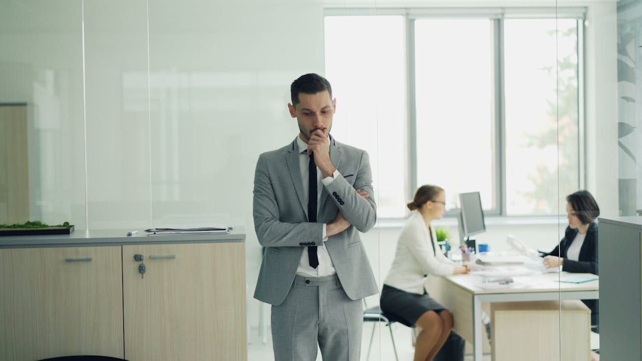 Worried business professional standing in office while colleagues work at desks, reflecting payroll stress and cash flow challenges.